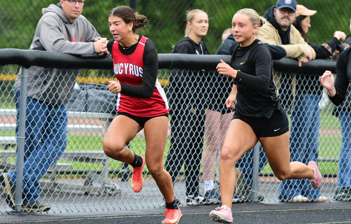 Two female runners sprinting along a track beside a chain-link fence as spectators watch from behind it.