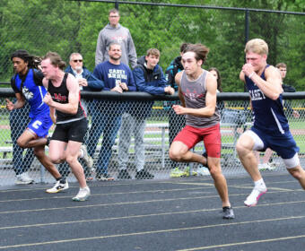 Four male sprinters dash on an outdoor track, with spectators watching from behind a chain-link fence.