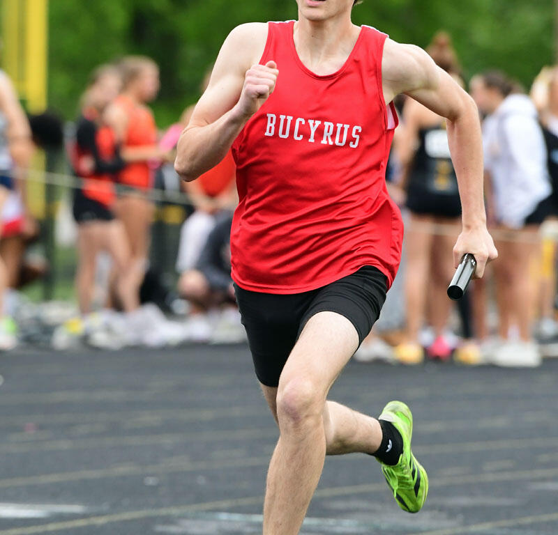 Male sprinter in a red Bucyrus jersey runs on a track holding a baton during a relay race, with onlookers in the background