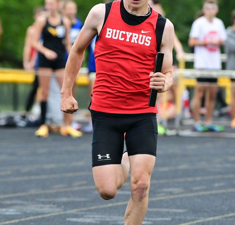 Male sprinter in a red Bucyrus singlet running on a track, holding a baton during a relay race.