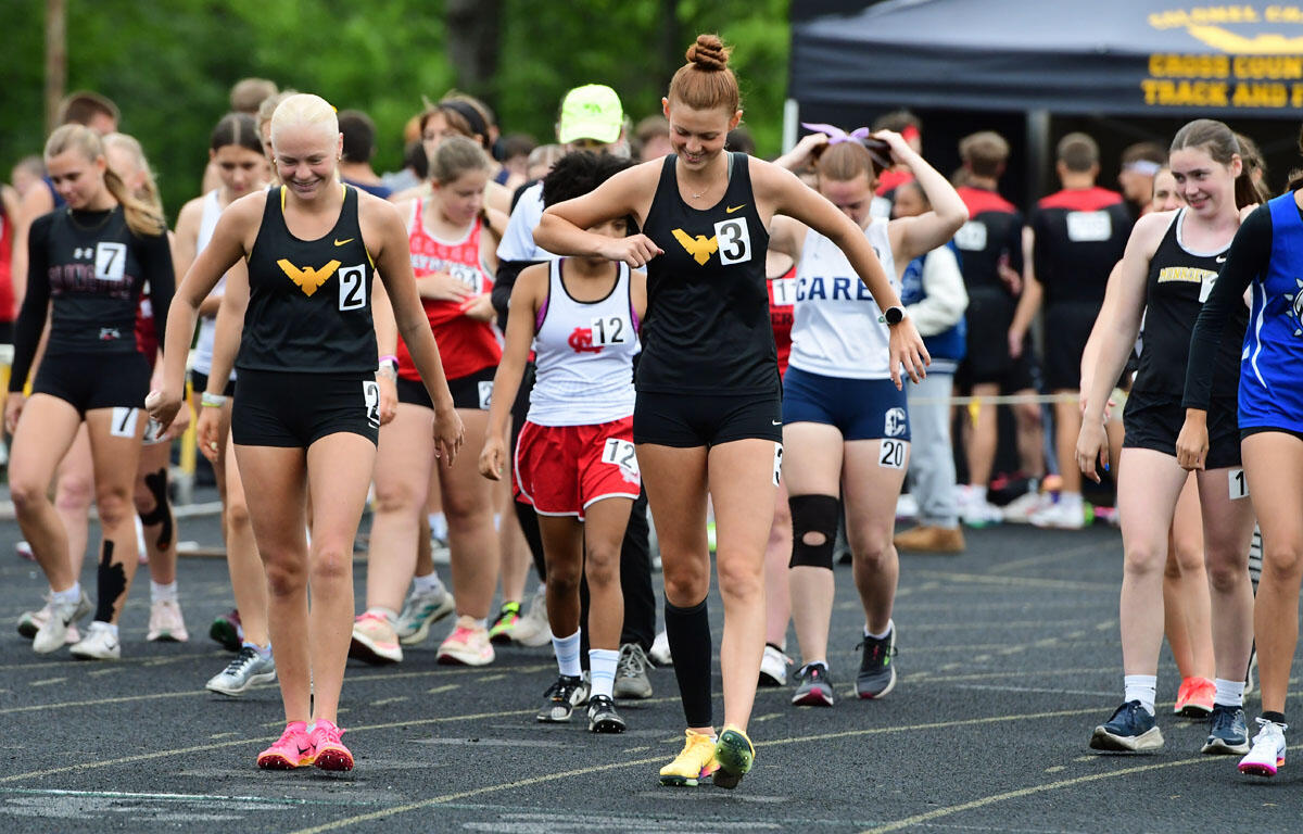Group of female sprinters at a track starting line, wearing numbered bibs and team uniforms, ready to race.