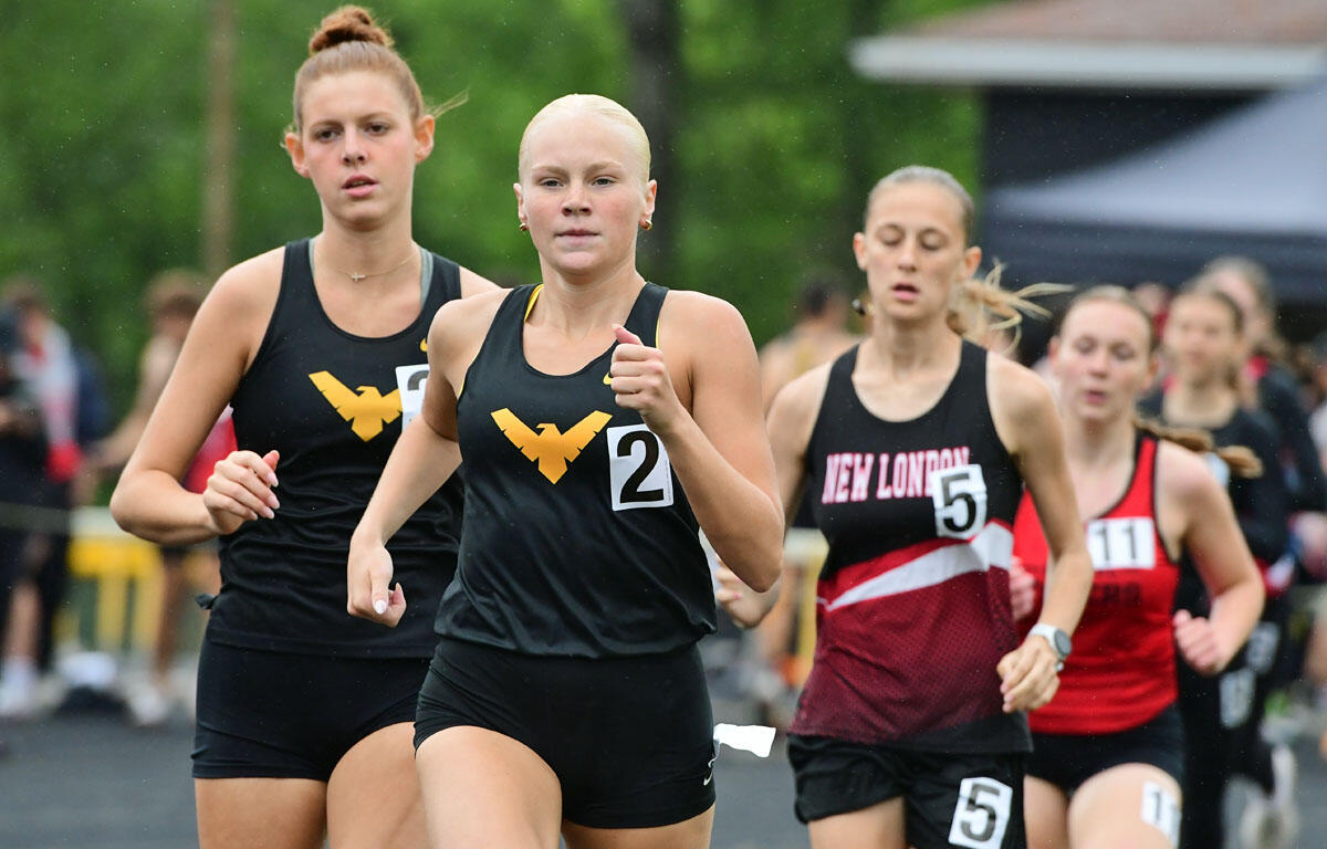 Female sprinter with shaved head leading a track race, wearing black singlet with gold wing logo; others in black and red uniforms behind him/her.