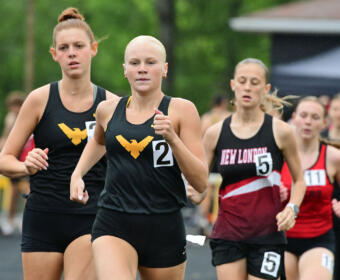 Female sprinter with shaved head leading a track race, wearing black singlet with gold wing logo; others in black and red uniforms behind him/her.