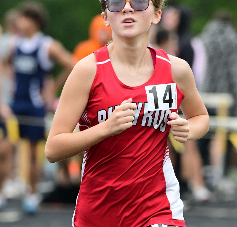 Young female runner in a red racing singlet with bib number 14, sprinting on an outdoor track with sunglasses on.