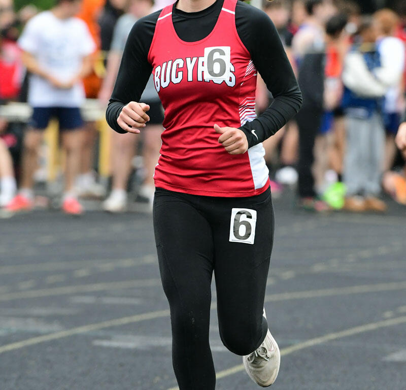Female runner wearing a red jersey with number 6, sprinting on a track with blurred spectators in the background.