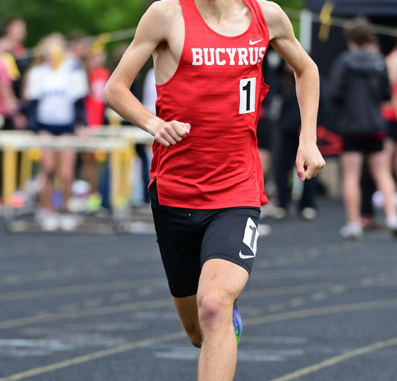 Young male runner in a red Bucyrus singlet sprinting on a track with race number 1 visible.