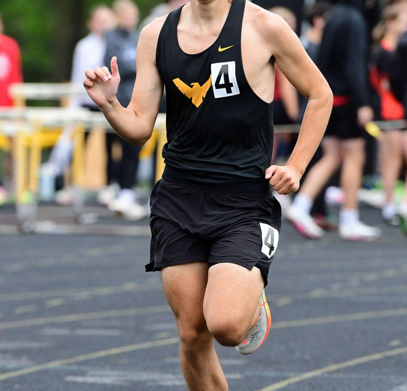 Male runner in a black singlet and shorts with bib 4, mid-race on a track Badges blurred spectators in background.