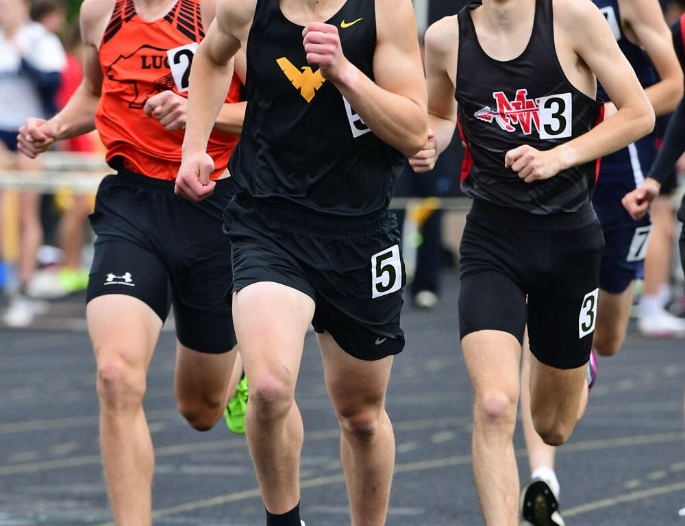Four young male runners sprinting on a track during a race; the front runner wears a black kit with a yellow emblem and leads the pack.