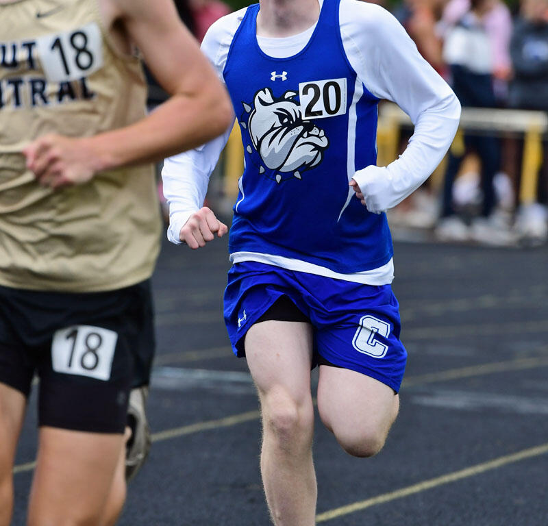 Young male runner in a blue singlet with number 20 sprinting on a track, spectators blurred in the background.