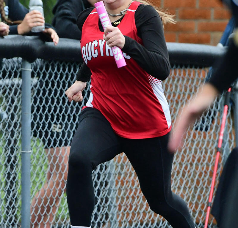 Female relay runner in a red jersey and glasses sprinting with a pink baton during a track race by a chain-link fence.