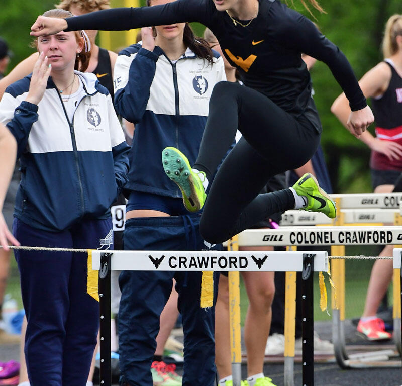 Female athlete in black athletic wear leaps over a hurdle on a track as teammates watch nearby.