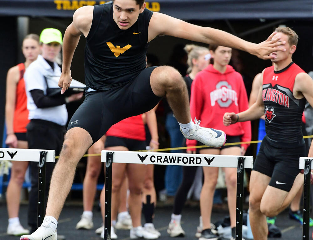 Male hurdler in a black singlet clears a hurdle mid-jump during a track meet; spectators and runners in the background.
