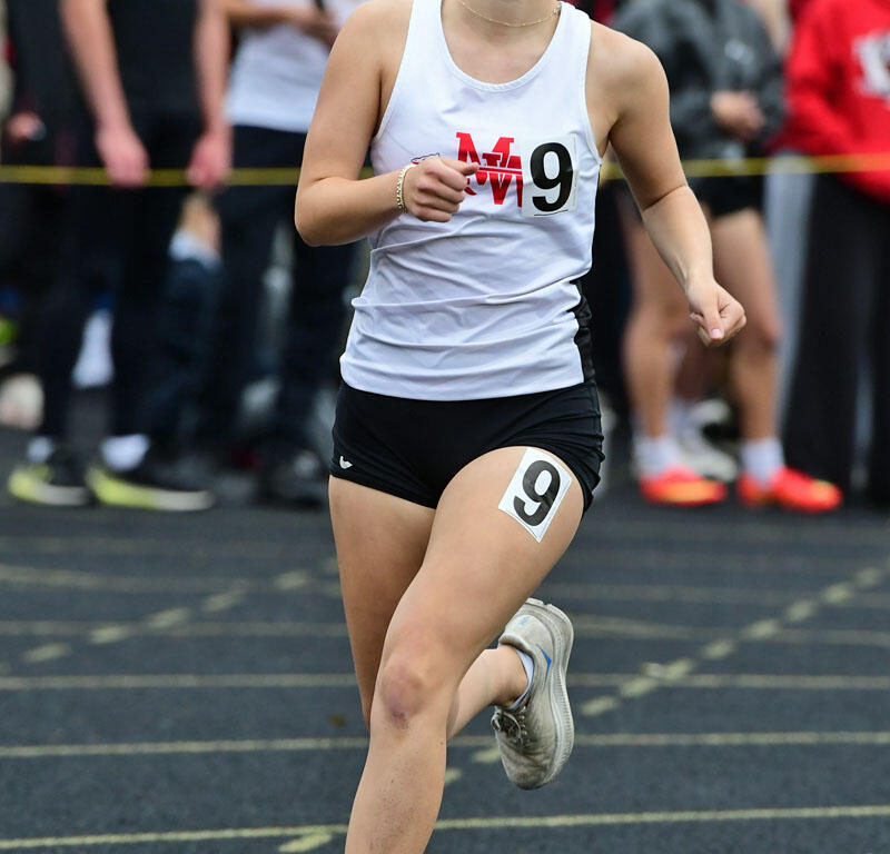 Female athlete sprinting on a track, wearing a white tank top with the number 9 and black shorts, mid-stride near an orange cone.