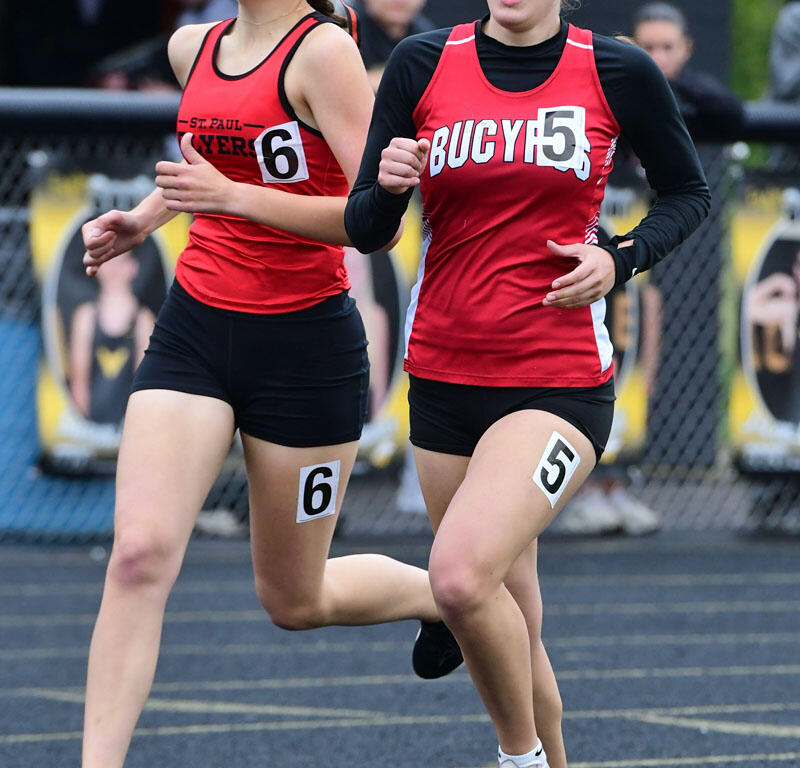 Two female track athletes in red uniforms race side by side on a blue track wear numbers 6 and 5.