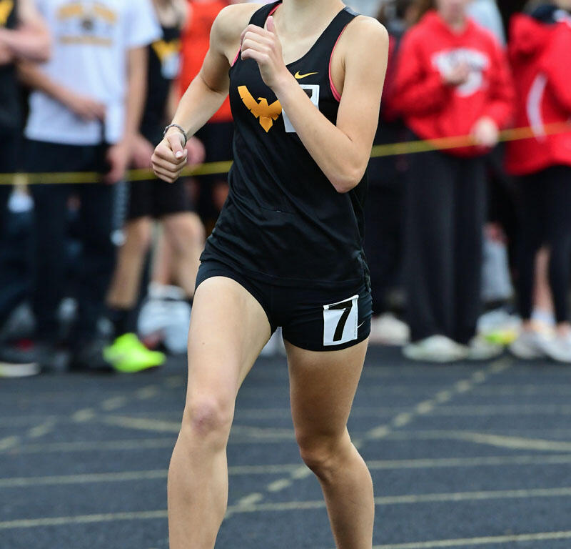 Female athlete in black uniform running on track during race, number 7 on shorts, with cheering crowd in background and an orange cone nearby.
