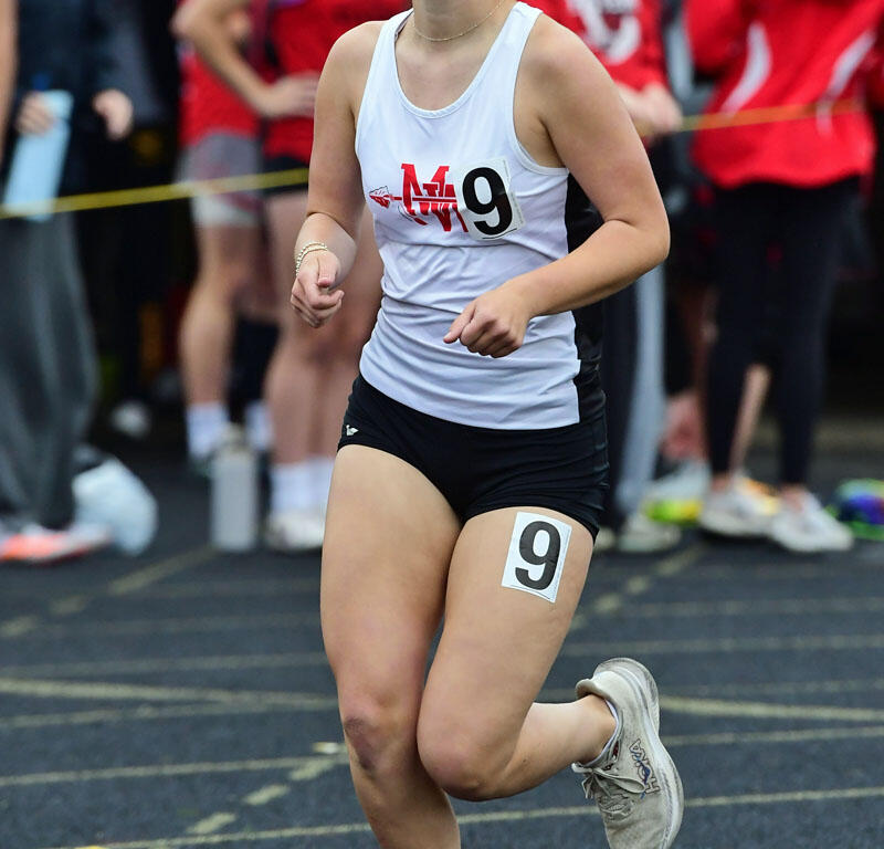Female track athlete in a white tank top and black shorts, sprinting on a blue track with bib number 9, against a backdrop of spectators and other runners.