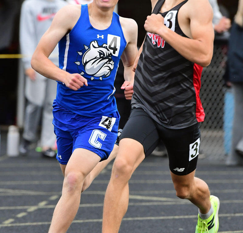 Two male sprinters race side by side on a track, one in blue with a bulldog logo and bib 4, the other in black with bib 3, mid-sprint.