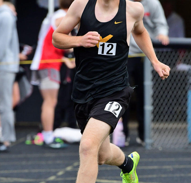 Young male sprinter in a black singlet with race bib 12, mid-run on a track with neon green shoes.
