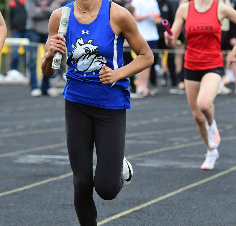 Female sprinter in a blue jersey runs with a baton during a relay race on a track, with a red-team competitor and spectators in the background.