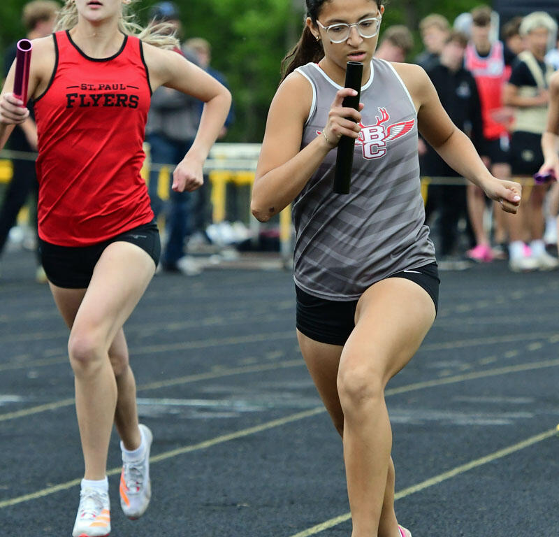 Two female athletes sprint on a track in a relay race: the foreground runner wears a gray top with pink shoes and holds a baton, while a teammate in a red St. Paul Flyers uniform follows behind.