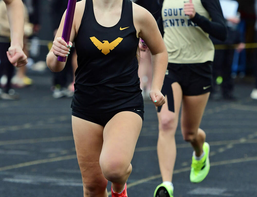 Female sprinter in a black singlet with a gold WVU logo runs on a track, gripping a purple relay baton as she advances; another runner in a beige jersey follows with spectators in the background.