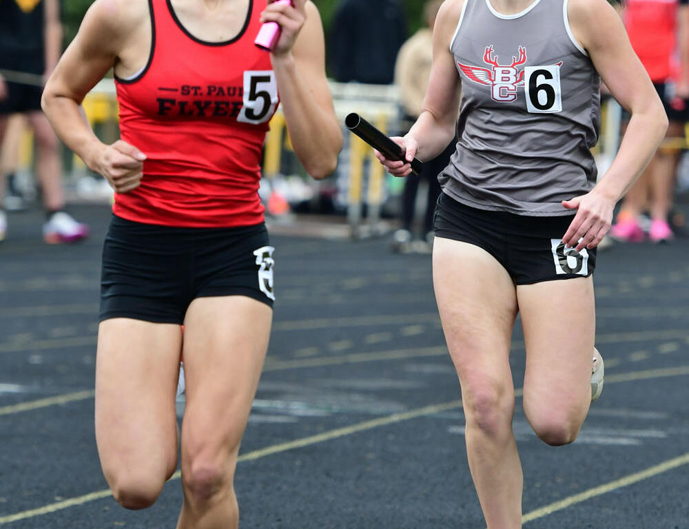 Two female sprinters in a relay race on a track; red uniform #5 carries a pink baton, ahead of gray uniform #6 with a black baton.