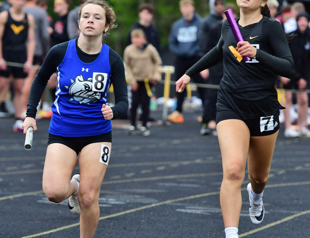 Two female sprinters in a relay on a track: left runner in blue shirt with number 8 holding a baton, right runner in black with number 7 and a purple baton, crowd in background.