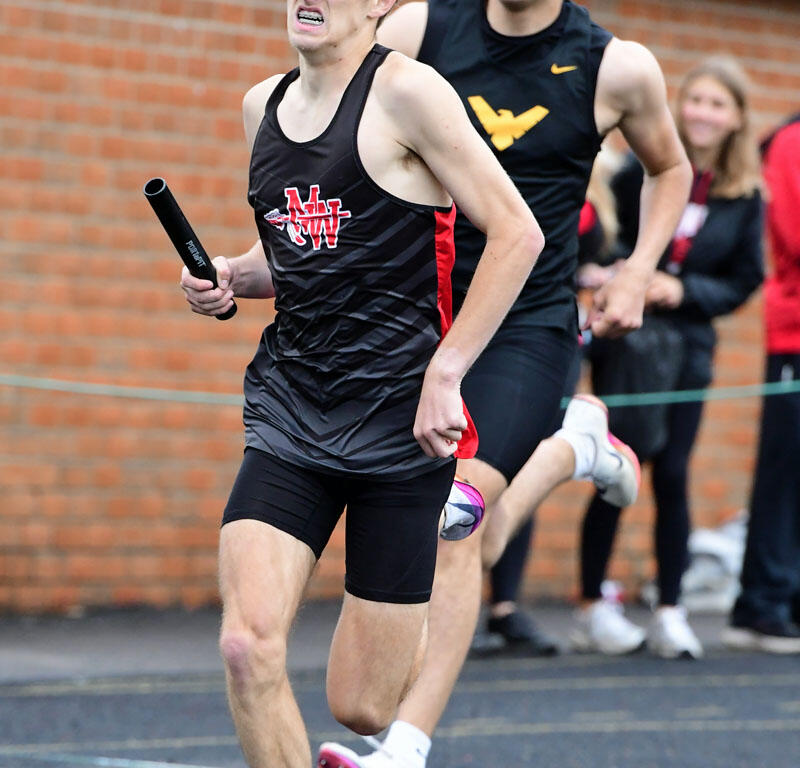 Relays race scene: two male sprinters running on a track, front runner in black-red uniform gripping a baton, hair flying, brick wall in background.