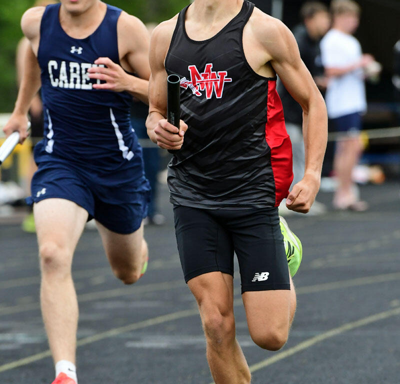 Two male sprinters race on a track; the front runner in a black-red NW jersey holds a baton while the other pursues behind him in a blue uniform.
