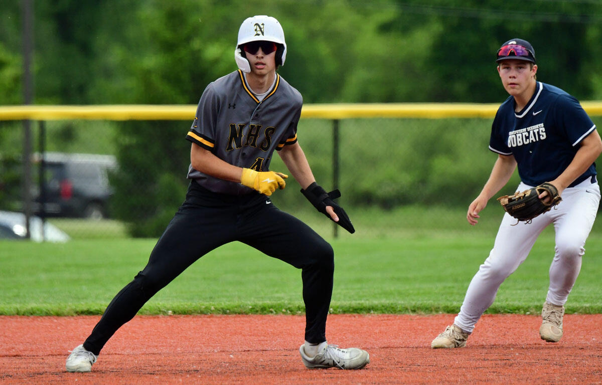 Baseball players in action on a field: a runner in a gray NHS uniform with helmet and yellow glove in a ready stance on the dirt, while a teammate in a navy uniform watches with a glove nearby.