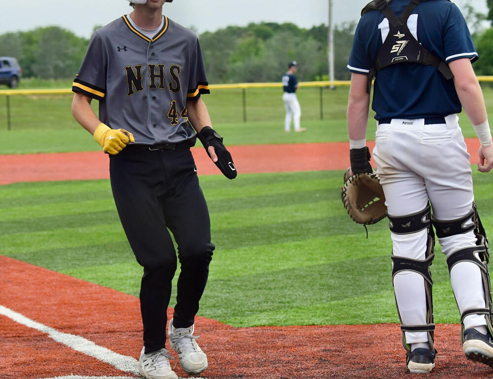 Two teenage baseball players stand near home plate on a field: one in a gray NHS uniform with helmet and yellow glove, the other in catcher's gear facing away.