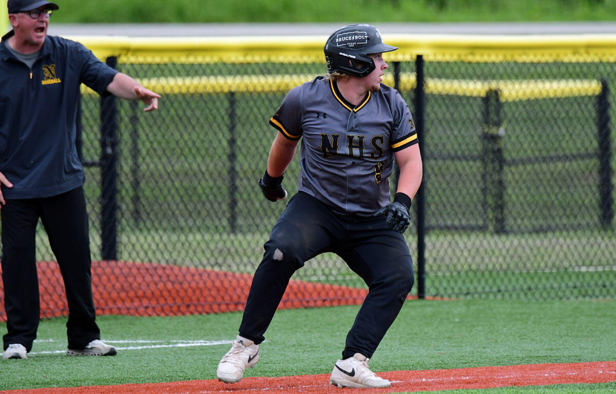 Baseball player in gray NHS uniform and helmet crouches and runs on the orange baselines while a coach points from the left.