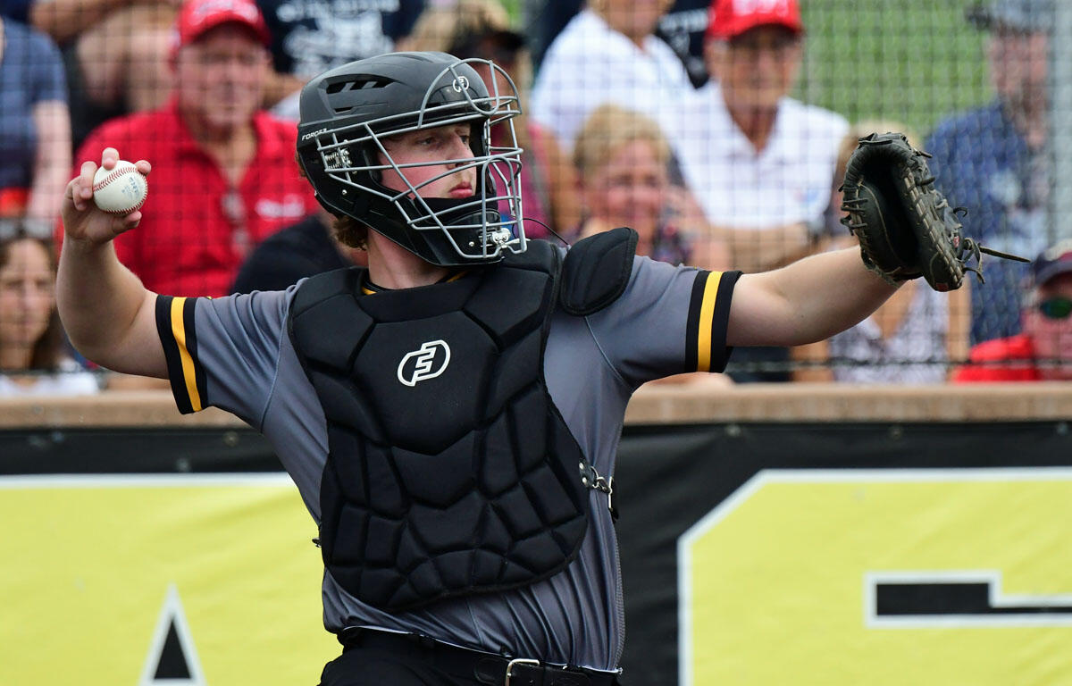 Baseball catcher mid-throw, wearing helmet and chest protector, in gray and black gear with a glove on the right hand and a ball in the left hand in a crowded stadium.