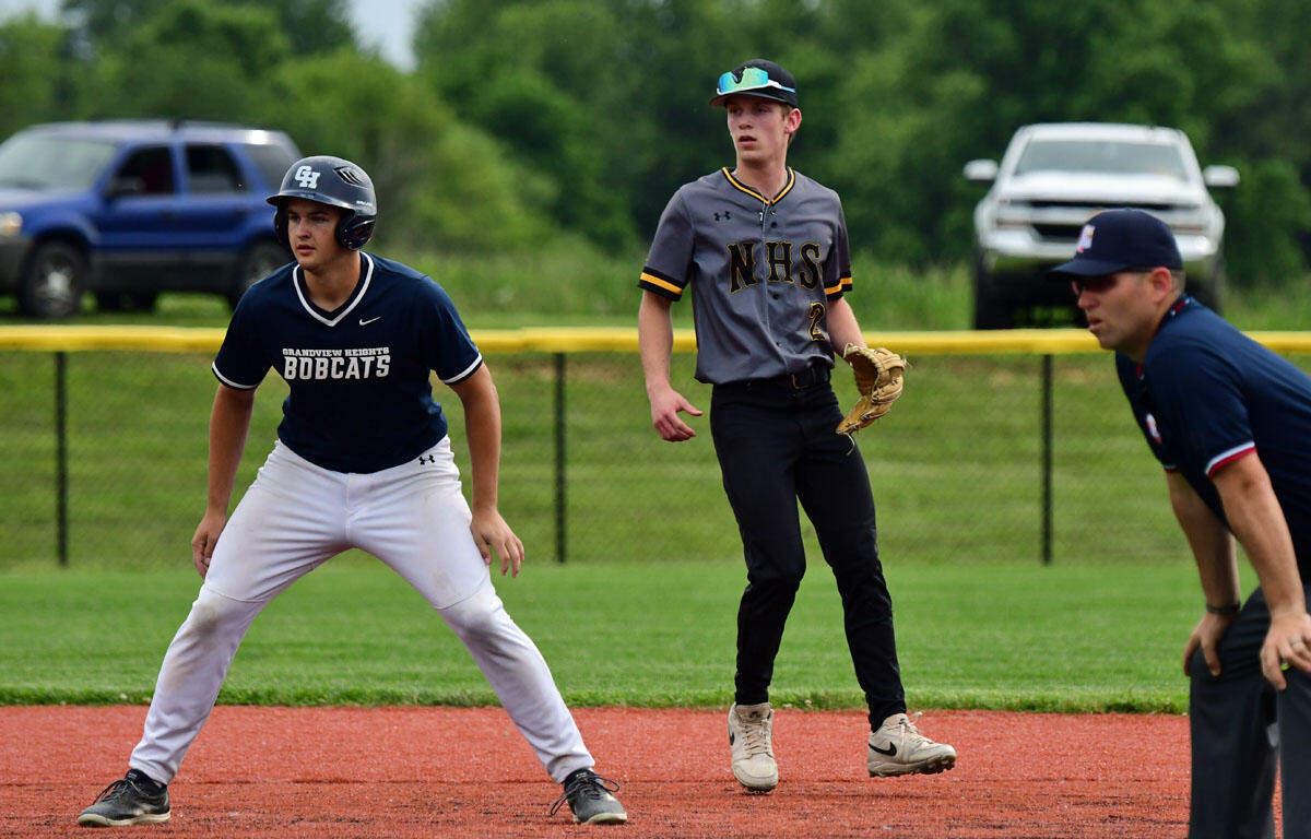 Baseball players on a field: runner in gray NHS jersey ready to field; infielder in navy Bobcats stance; coach or umpire on right watching.