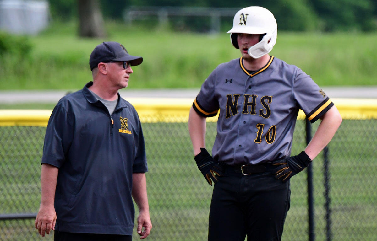 Baseball coach talks with a player in a gray NHS uniform and white helmet near the outfield fence, #10 on the jersey.