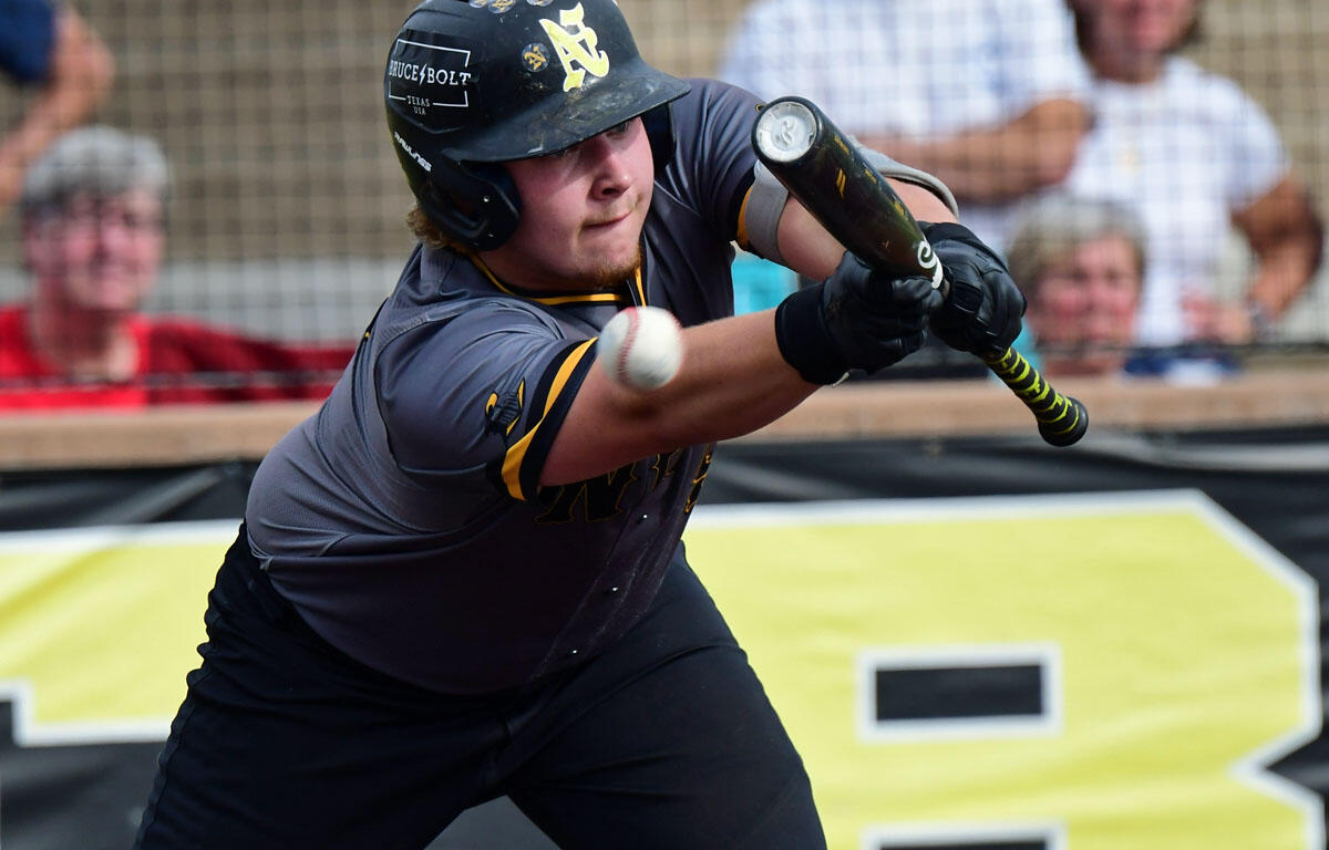 Baseball batter in a gray and yellow uniform swings at a pitched ball, with spectators in the background behind the fence.