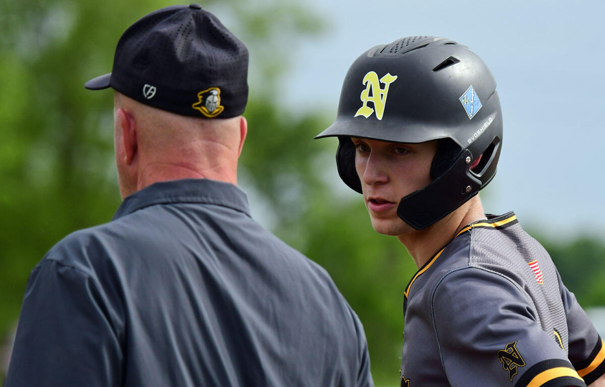 Baseball player in a gray uniform with a protective helmet talking to a coach seen from behind outdoors on a sunny day
