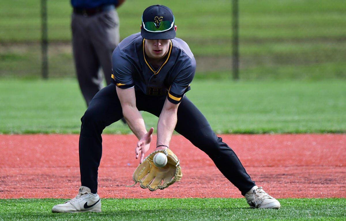 Baseball infielder crouches to field a ground ball, glove low to the dirt in a gray and navy uniform with a cap and sunglasses on the brim.