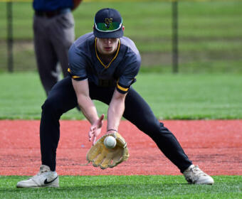 Baseball infielder crouches to field a ground ball, glove low to the dirt in a gray and navy uniform with a cap and sunglasses on the brim.