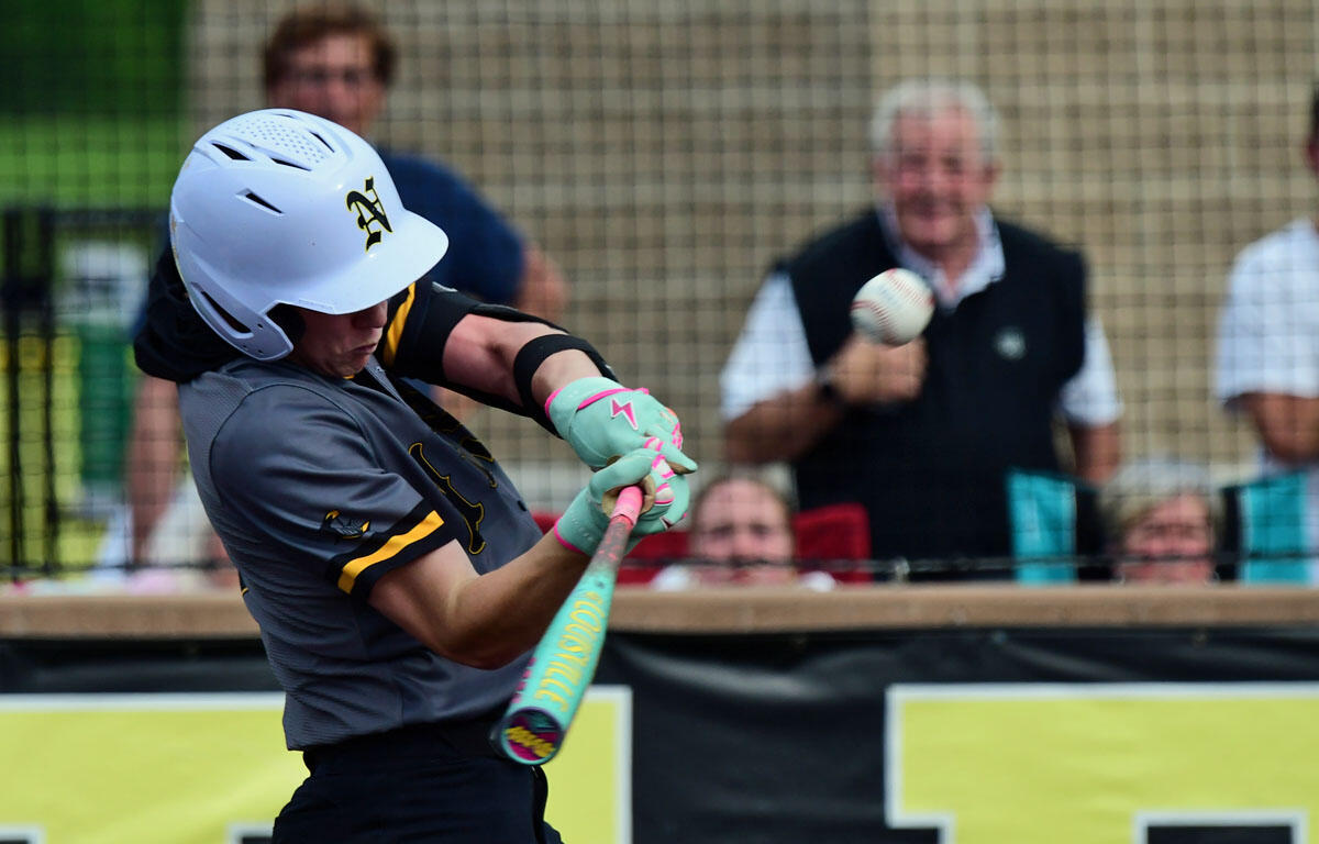 Female softball player wearing a helmet and teal gloves swings a bat during a game, with spectators behind a fence in the background.
