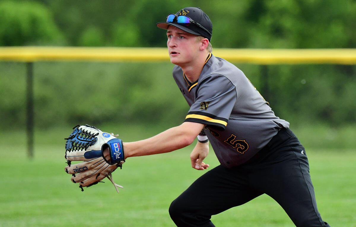 Baseball player in a gray uniform and black cap extends a blue glove to field a ball on a grassy field.