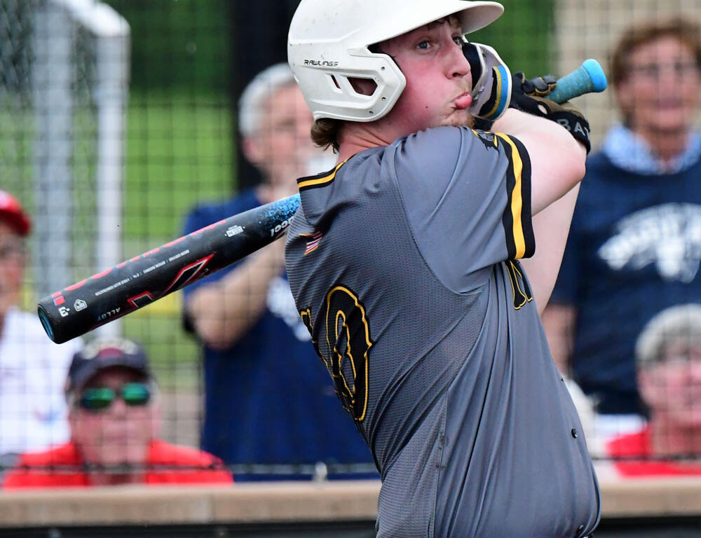 Baseball player in a gray jersey and white helmet swings a bat during a game, with spectators in the background.