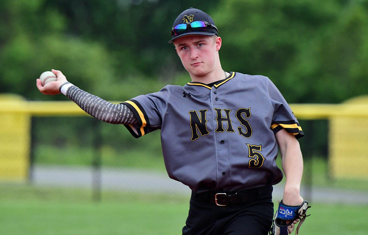 Young baseball pitcher in a gray NHS jersey throws a ball on a grassy field during a game.