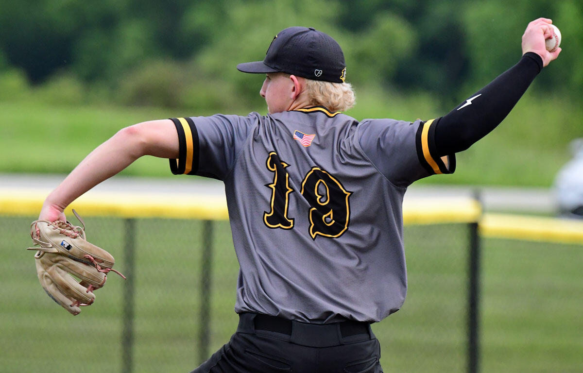 Baseball pitcher in gray jersey with number 19 throws a pitch, glove in left hand and ball in right, on a field with a chain-link fence behind.