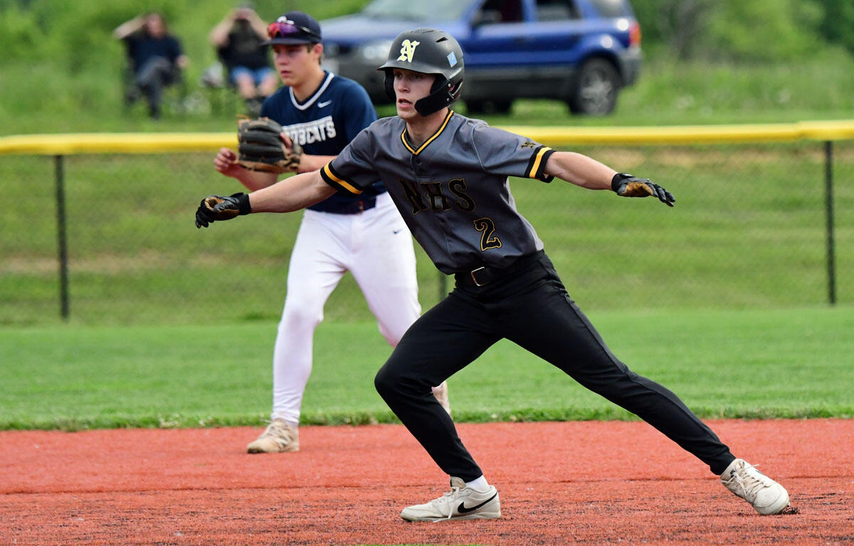 Baseball fielder in a gray NHS uniform and helmet stretches for a ground ball on the infield; teammate in navy watches from behind on the grass.