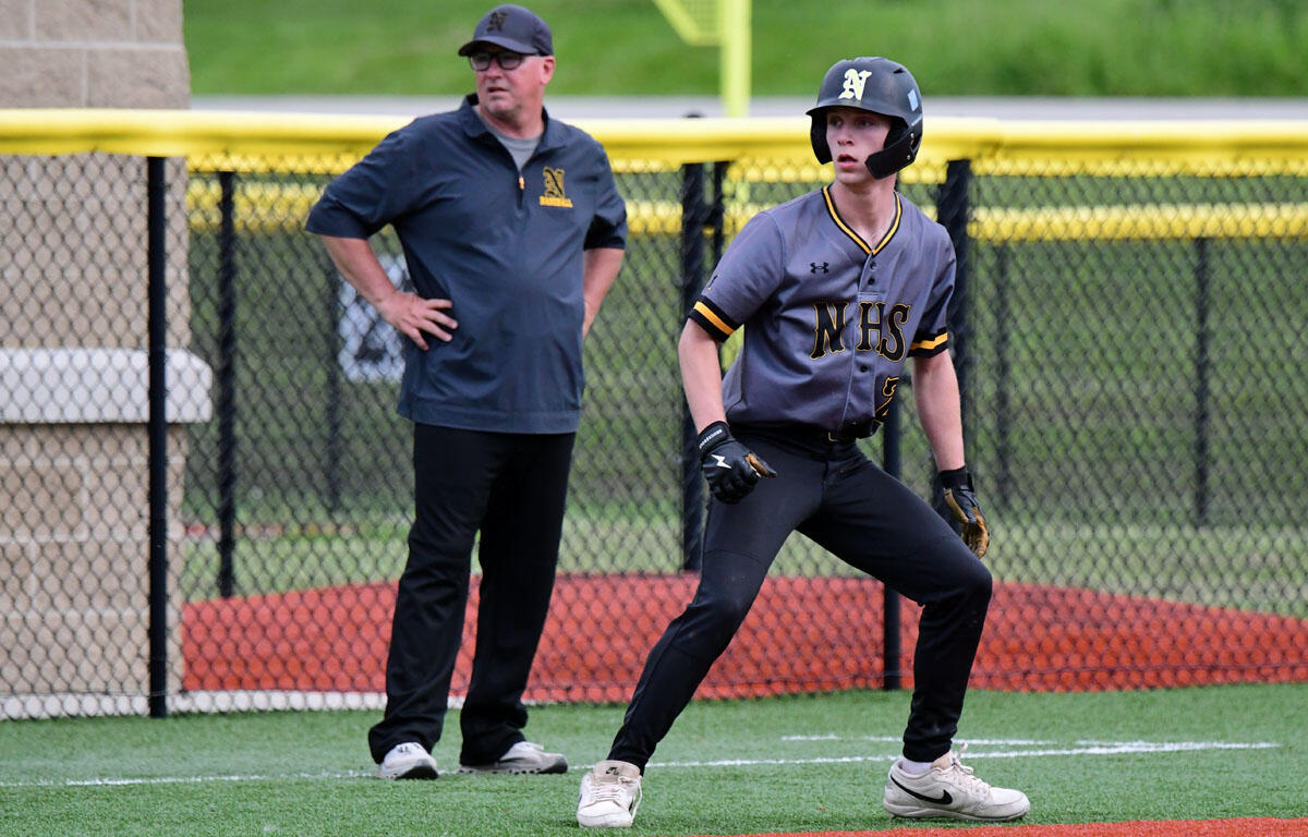 Young baseball player in a gray NHS uniform with a helmet, ready to run, as a coach watches from behind the fence.