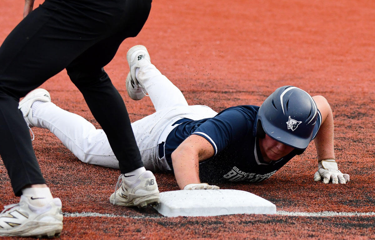 Baseball player slides into a white base on a dirt field as another player looms nearby.
