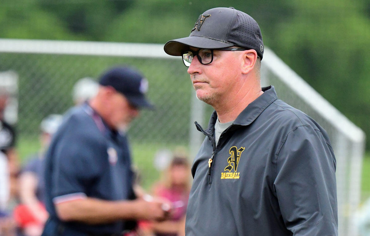 Baseball coach in a gray cap and jacket, wearing glasses, standing on a field with a blurred crowd in the background.