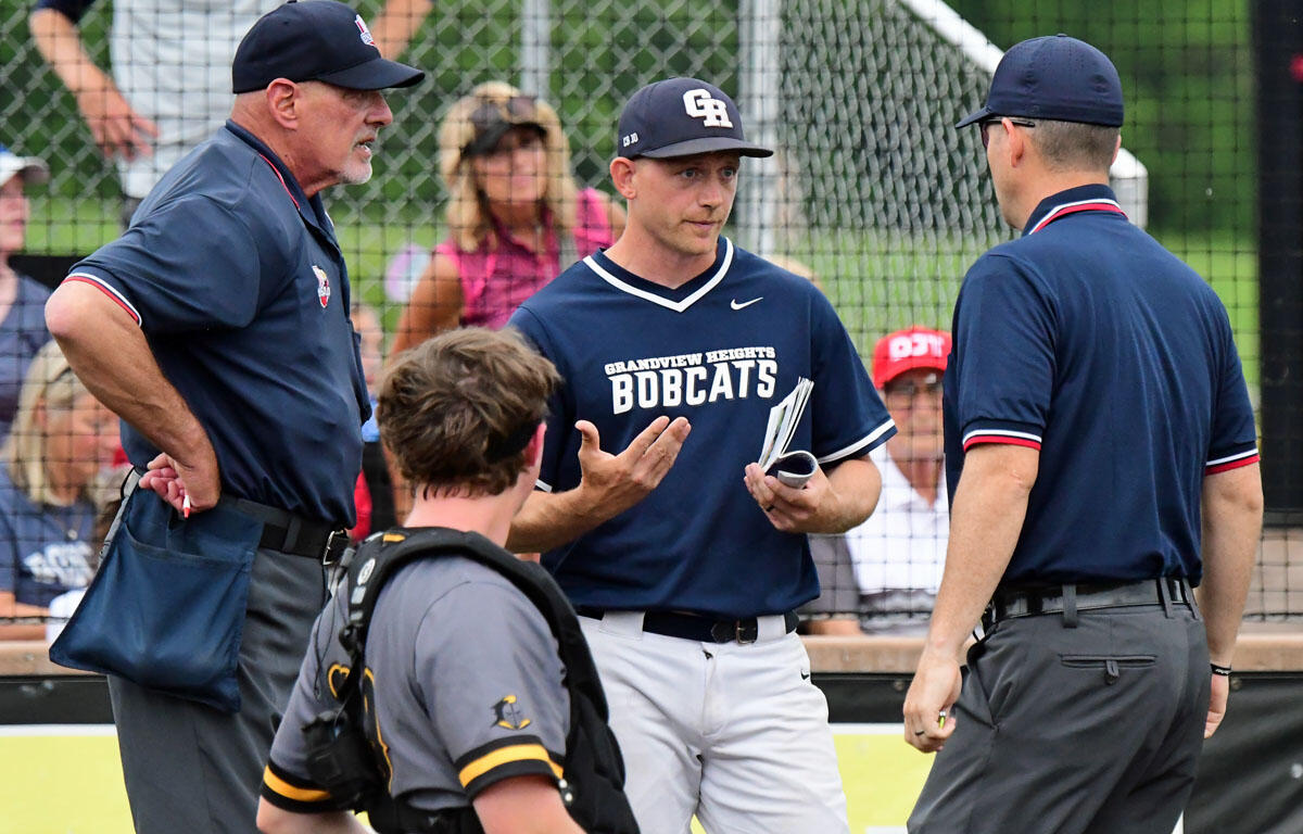 Baseball coach in dark blue shirt talking to players near the sideline, with a catcher in gear and another official nearby.