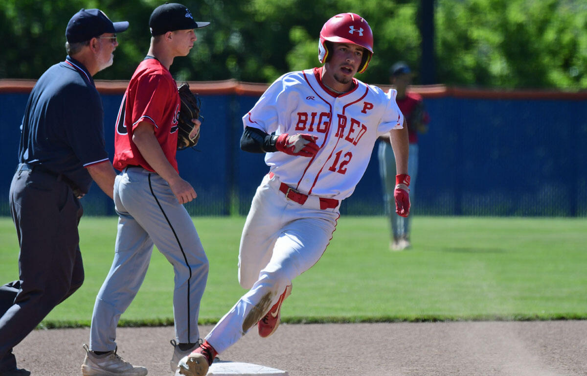 Baseball player in a white and red 'BIG RED' jersey slides into first base as a coach/teammate in red and an umpire watch nearby on the infield dirt.